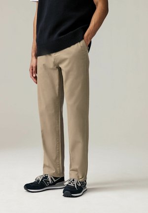 Beige gerade geschnittene Hose mit Seitentaschen, kombiniert mit schwarzen Sportschuhen mit weißen Schnürsenkeln und grauen Akzenten. Darüber ein gerippter schwarzer Pullover.