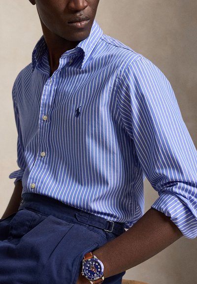 Άνδρας που φοράει μπλε και άσπρο ριγέ πουκάμισο με κουμπιά και γυρισμένα μανίκια, σκούρο μπλε παντελόνι και σκούρο μπλε ρολόι με καφέ δερμάτινο λουράκι.