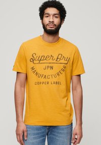 T-shirt di cotone gialla con collo rotondo, caratterizzata da un logo stampato scuro che recita "Super Dry JPN Manufacturers Copper Label" sul davanti.