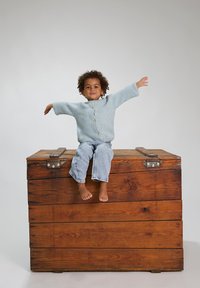 Cardigan lavorato a maglia blu chiaro con grandi bottoni, indossato sopra jeans in denim chiaro. Seduto su un baule in legno marrone, a piedi nudi con le braccia tese.