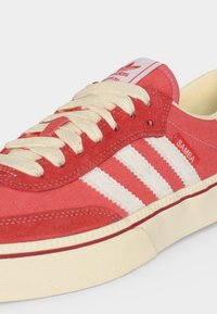 Κόκκινο αθλητικό παπούτσι Adidas Samba με λευκές ρίγες, εκρου κορδόνια και μπεζ σόλα, εμφανίζοντας πλευρικές και κορυφαίες λεπτομέρειες.