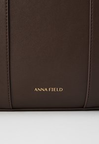 Tamnosmeđa kožna torba s vertikalnim šavovima i zlatnim ugraviranim logotipom "ANNA FIELD" blizu donjeg središnjeg dijela.