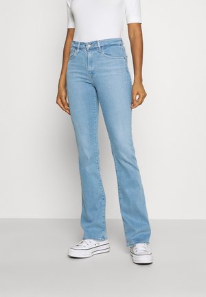 Personne portant un jean droit taille haute bleu clair, un haut blanc à manches courtes et des baskets compensées blanches, debout devant un fond uni.