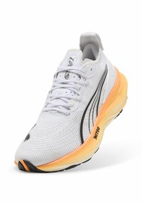 Zapatilla deportiva blanca con una parte superior de malla texturizada, acentos negros y una suela gruesa en naranja y crema. Presenta cordones planos y un collar acolchado.
