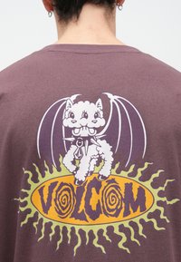 Άτομο που φορά καφέ πουκάμισο με εικόνα λευκού δίκερου φτερωτού σκύλου πάνω από κίτρινη έκρηξη ήλιου και το κείμενο "VOLCOM" σε στυλιζαρισμένη γραμματοσειρά.