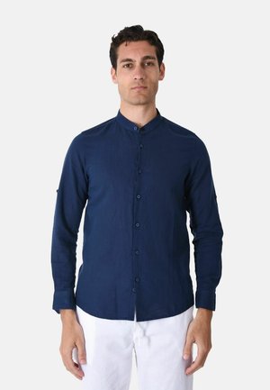 Uomo in piedi, indossa una camicia blu navy con collo alla coreana abbottonata, maniche lunghe e pantaloni bianchi su sfondo semplice.