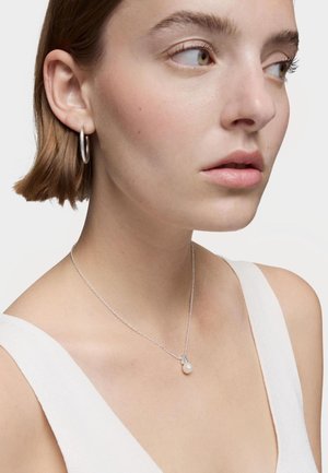Collar de cadena de plata con un pequeño colgante de perla. La modelo lleva una blusa sin mangas, mostrando la joyería sobre su piel clara.