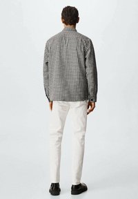 Homme portant une veste pied-de-poule noire et blanche, un pantalon blanc et des chaussures noires, debout de dos sur un fond uni.