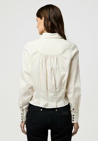 Blusa crema accorciata con pieghe posteriori, colletto classico e polsini abbottonati. Tessuto morbido con dettagli di trama appena percettibili. Abbinata a jeans neri.