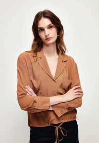 Braune Satinbluse mit Kragen, Knopfleiste, zwei Brusttaschen und tailliertem Bund. Sie hat lange Ärmel mit Knopfmanschetten.