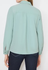 Femme portant une chemise à manches longues vert clair et un jean bleu foncé, vue de dos sur un fond blanc.