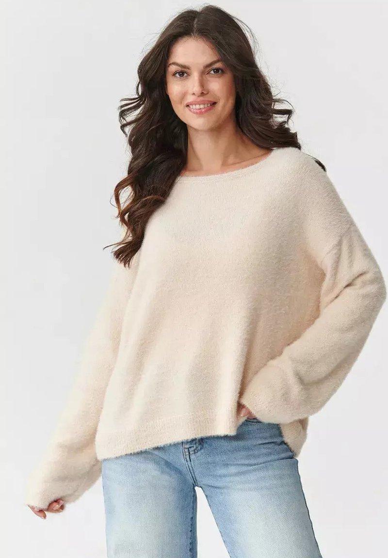 Jasno beżowy, oversize sweter o miękkiej, puszystej teksturze, z szerokim dekoltem w kształcie łodzi i długimi rękawami. W połączeniu z jasnoniebieskimi dżinsami.