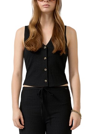 Femme aux longs cheveux châtain clair portant un haut noir sans manches à boutons et un pantalon noir assorti avec cordon de serrage, bracelet doré au poignet.