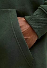Sudadera verde olivo con superficie texturizada, que presenta un bolsillo lateral y puños acanalados, mostrando una mano en el bolsillo para dar perspectiva.