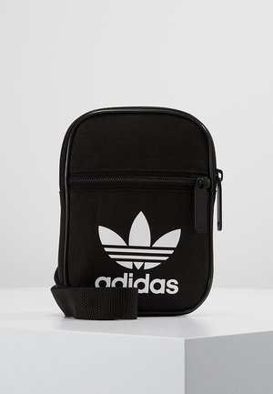 Musta Adidas-läpälaukku valkoisella kolmilehtilogolla, etutaskulla, jonka vetoketju, ja säädettävällä kudotulla olkahihnalla vaalealla yksivärisellä taustalla.