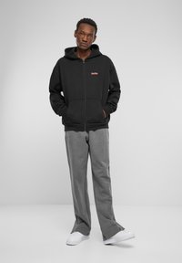 Crna zip-up hoodie s kapuljačom i prednjim džepovima, s malim crvenim logom. Uparena sa svijetlo sivim hlačama i bijelim tenisicama.