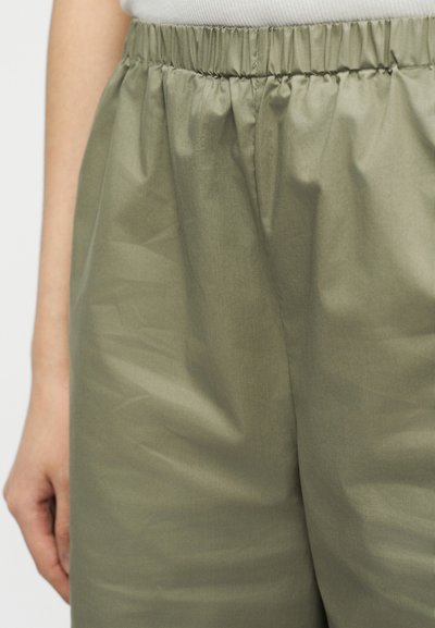 Κοντινό πλάνο ελαστικής μέσης σε παντελόνι ανοιχτού πράσινου ελιάς, φορεμένο με λευκή μπλούζα, που δείχνει μέρος του χεριού και του βραχίονα του φορέα.