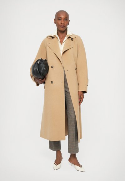 Trench coat din lâni bej cu nasturi dubli, purtat peste o cămașă deschisă la culoare și pantaloni în carouri. Geantă neagră și pantofi cu vârful ascuțit de culoare crem.