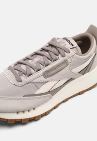 Baskets Reebok gris clair et blanc avec semelle texturée, devant à lacets, panneaux en suède et tissu, et étiquettes de marque sur le côté et la languette.