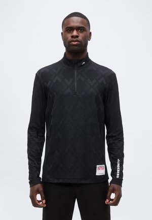Homme debout portant un haut noir à manches longues zippé avec des motifs géométriques subtils, logo blanc sur la manche et étiquette près de la taille.