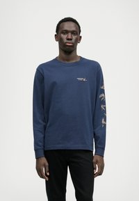 Tumesinise pikakäistega T-särk, mis on valmistatud puuvillast ja millel on ümar kaelus. Esiküljel on väike logo ja värvilised linnugraafikud pikki varrukaid.