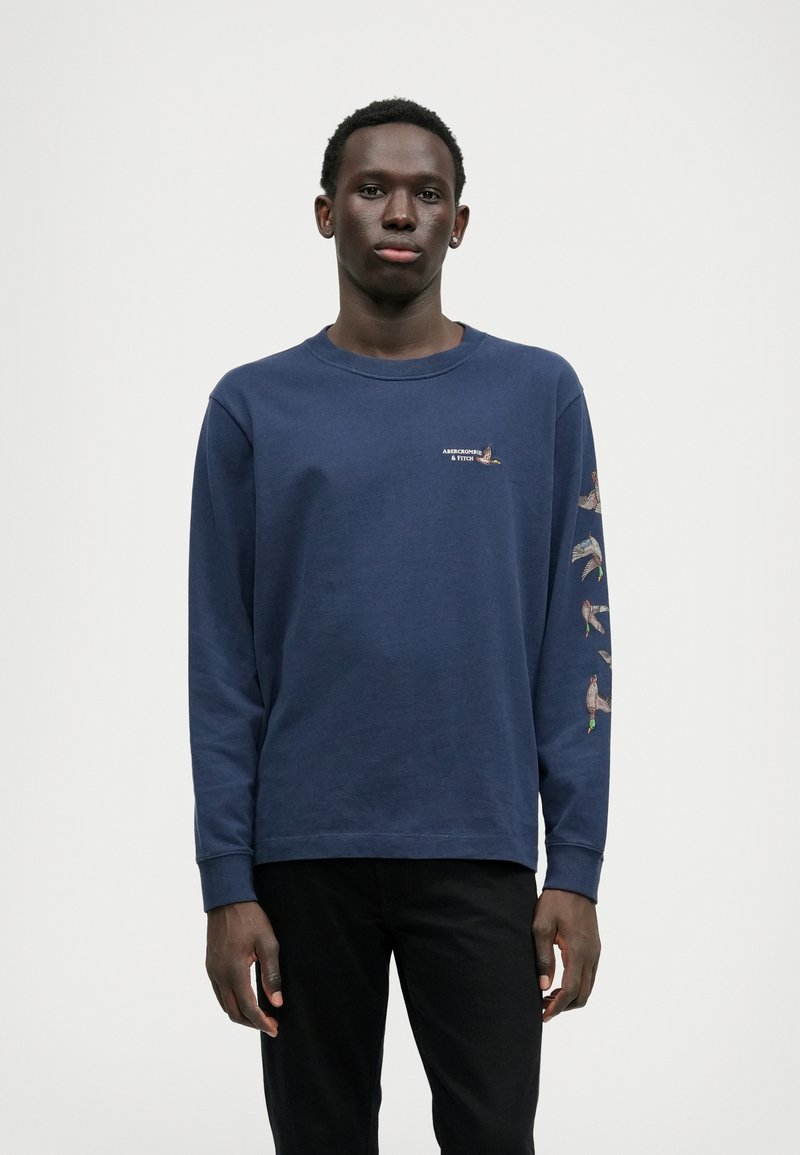 Tumesinise pikakäistega T-särk, mis on valmistatud puuvillast ja millel on ümar kaelus. Esiküljel on väike logo ja värvilised linnugraafikud pikki varrukaid.