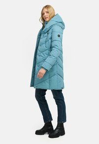 Jasnoniebieska kurtka puchowa z kapturem, z pikowanym wzorem, zapinana na napy i z bocznymi kieszeniami, zestawiona z ciemnymi dżinsami i czarnymi butami.