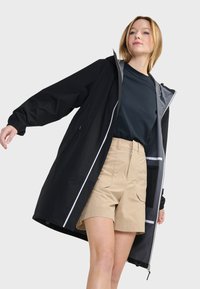 Giacca impermeabile nera con cappuccio, foderata in grigio e con dettagli riflettenti. Indossata sopra una maglietta blu e pantaloni corti beige, con tasche laterali.