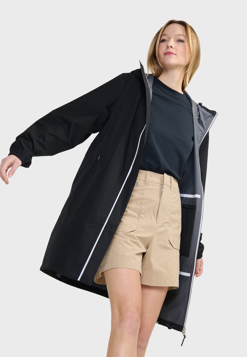 Giacca impermeabile nera con cappuccio, foderata in grigio e con dettagli riflettenti. Indossata sopra una maglietta blu e pantaloni corti beige, con tasche laterali.