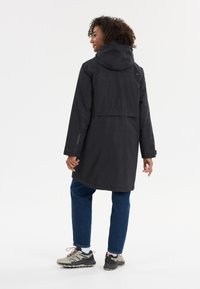 Giacca impermeabile nera con cappuccio, caratterizzata da una vestibilità comoda, maniche lunghe e un design con schiena spaccata. Abbinata a jeans blu e sneaker.