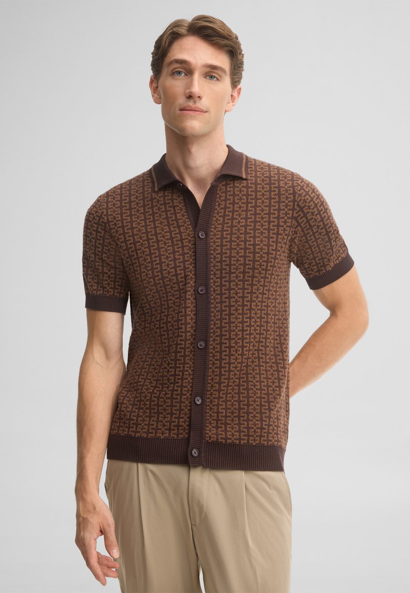 Man wearing a brown short-sleeve button-up shirt with a geometric pattern and beige pants, standing with one arm behind his back.