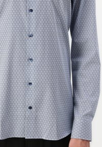 Lys blå skjorte med mønster som har et geometrisk design, med mørke knapper og krage med knapper. Lange ermer avsluttes med mansjetter.