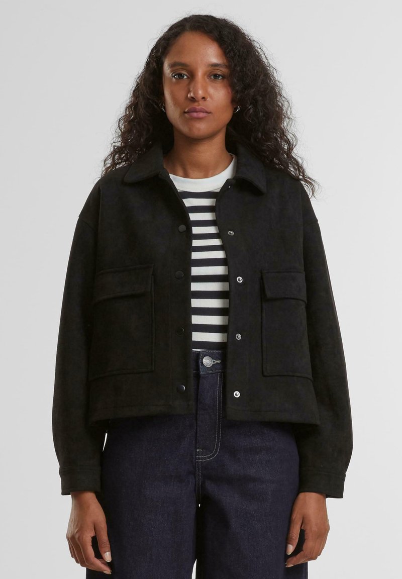 Žena s kudrnatými vlasy, která má na sobě černou zapínací bundu přes černobíle pruhované tričko a tmavě modré džíny s vysokým pasem.