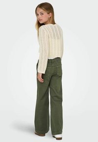 Biely pletený krátky sveter s dierovaným vzorom a dlhými rukávmi v kombinácii s vysokými olivovo zelenými nohavicami širokého strihu a bielymi teniskami.