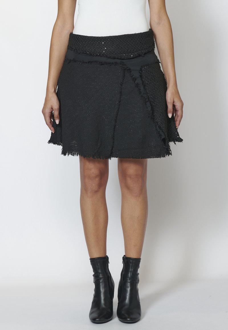 Femme portant une jupe noire texturée en ligne A avec des bords effilochés et des bottines noires, debout devant un fond clair uni.