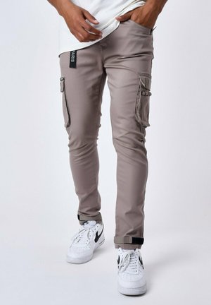 Pantalones cargo beige con un ajuste slim, que cuentan con bolsillos laterales, correas en los tobillos y una etiqueta de marca negra en el lado izquierdo. Combinados con zapatillas blancas.