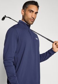 Μπλε ναυτικό, μακρυμάνικο πουλόβερ με φερμουάρ στον γιακά και λογότυπο Under Armour στο στήθος; ελαφρύ ύφασμα; γκολφ μπαστούνι κρατημένο πάνω στον ώμο.