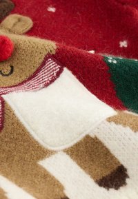 Couverture en tricot présentant un motif de renne en beige et rouge avec des accents blancs, des détails verts et une texture douce et pelucheuse.