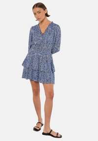 Blau gemustertes Kleid mit langen Ärmeln, V-Ausschnitt, Rüschenschichten und lässigem Schnitt. Kombiniert mit schwarzen Riemchensandalen.