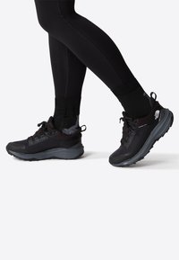 Scarpe da atletica nere con una tomaia texturizzata, lacci sicuri e una suola spessa. Indossate con leggings e calze nere, evidenziando un design sportivo.