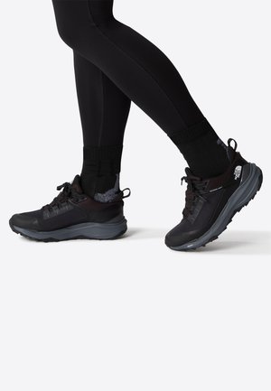 Scarpe da atletica nere con una tomaia texturizzata, lacci sicuri e una suola spessa. Indossate con leggings e calze nere, evidenziando un design sportivo.