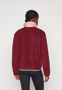 Person med flettehår iført en bordeaux fleecejakke med høy rosa krage og svarte leggings, vendt bort fra kameraet mot en ensfarget bakgrunn.