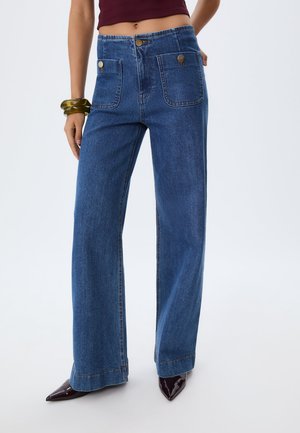 Blaue, weit geschnittene Jeans haben eine hohe Taille, zwei vordere Taschen mit Knopfverschluss und eine glatte Denim-Oberfläche. Dazu werden schwarze, spitz zulaufende Schuhe getragen.