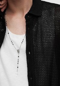 Maglietta nera in maglia sopra una T-shirt bianca, con una collana in argento che presenta perline nere e un pendente a croce bianco.