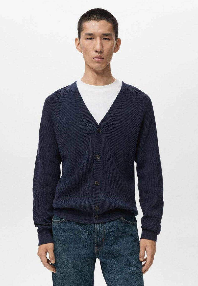 Cárdigan de punto de color navy con escote en V, cierre de botones y mangas largas. Llevado sobre una camiseta blanca de cuello redondo, combinado con jeans azules.