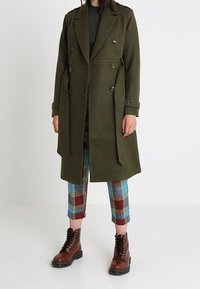 Frau trägt einen langen olivgrünen, zweireihigen Mantel, bäuerliche blaue und rote Karottenhose sowie braune Schnürstiefeletten.