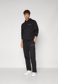 Hoodie noir et pantalon de survêtement en mélange de coton, avec une poche avant, broderie de logo, capuche avec cordon de serrage, et baskets blanches.