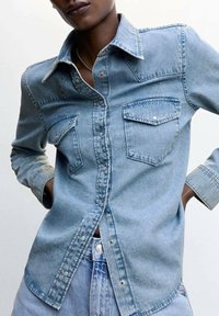 Helles blaues Jeanshemd mit klassischem Kragen, langen Ärmeln, zwei Brusttaschen und Druckknopfverschluss, das über eine leicht verblasste Textur verfügt.