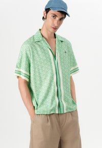 Short-sleeve green patterned shirt featuring a geometric design, light buttons, and striped accents. Paired with brown shorts and a denim cap.