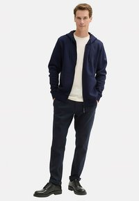 Sweat à capuche zippé bleu marine avec cordon de serrage, poches kangourou, porté sur un t-shirt blanc, associé à un pantalon foncé et des chaussures noires à lacets.